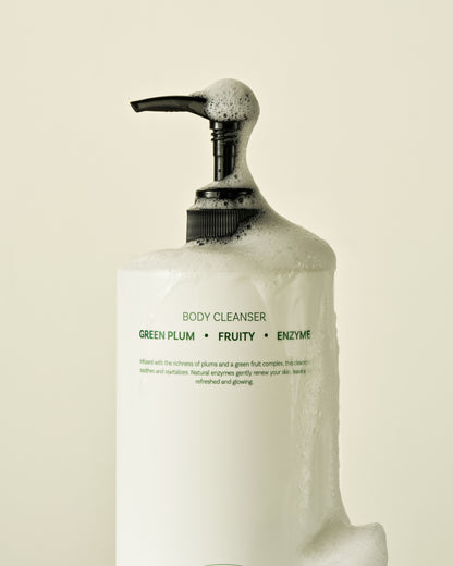 Close-up of green plums and papaya fruit next to the Green Plum Fruit Enzyme Body Cleanser bottle, highlighting the natural ingredients.